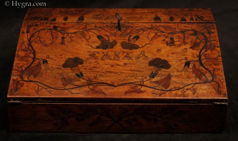The Eliza writing slope: an unique satinwood writing slope with both fine marquetry and inlay depicting  trailing convolvulus. ( wikipedia.org/wiki/Convolvulus  ) This is an artwork of love. The top is solid satinwood and inlaid. the sides have a saw-cut veneer of about 1mm. The accuracy of the marquetry is truly skillful.  On the front there is   a depiction of oak branches laden with acorns crossed.  The slope opens down to a velvet (replacement) writing surface and the usual compartments for inkwells and pens. Under the writing surface there is the usual place for paper storage and a nest of  three drawers  of part dovetail construction. This is not a manufactured piece, but rather a work of love by a now anonymous craftsman for his beloved Eliza who was clearly interested in flora.  Circa  1830.   Enlarge Picture
